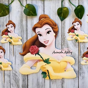 May include: A collection of Belle-themed cupcake toppers. The toppers feature Belle in her yellow gown, holding a red rose. The background is a light wood grain. Green leaves and vines are at the top. The name "Amanda Ashley" is written on the image.
