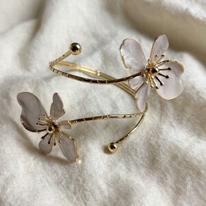 May include: A gold-tone metal cuff bracelet featuring a spiral design with two light pink, pearlescent flower-shaped accents at each end. The band has a textured, ridged surface and terminates in small, smooth gold-tone spheres at both ends.