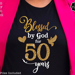 May include: A black t-shirt with gold glitter lettering that reads "Blessed by God for 50 years". A gold glitter heart and a gold glitter dove are on either side of the number 50.