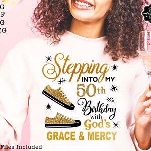 May include: A white sweatshirt with gold glitter lettering that says "Stepping into my 50th Birthday with God's Grace & Mercy". The text is surrounded by gold glitter stars and two gold glitter trainers.
