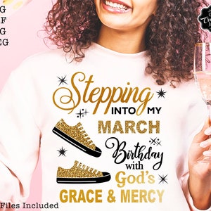 Może przedstawiać: Biała bluza z napisem w kolorze złotego brokatu: "Stepping into my March Birthday with God's Grace & Mercy". Na przodzie bluzy znajdują się dwie złote brokatowe trampki.