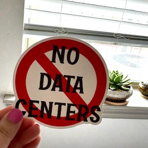 May include: A circular sticker featuring a white background with a red border and a diagonal red slash across the center. The text NO DATA CENTERS is printed in bold black capital letters across the front of the sticker design.