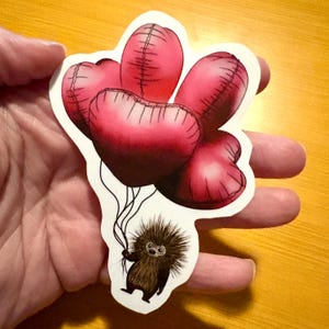 Puede incluir: Una pegatina con un puercoespín de dibujos animados sosteniendo un manojo de globos rojos en forma de corazón. Los globos tienen un diseño cosido y el puercoespín es marrón con un aspecto espinoso. La pegatina tiene un borde blanco.