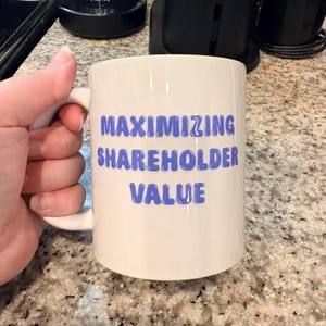 May include: A white ceramic mug with the text "MAXIMIZING SHAREHOLDER VALUE" in blue letters. The mug has a curved handle and is held by a hand. The background includes kitchen appliances and a speckled countertop.
