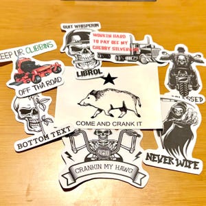 Pode incluir: Uma coleção de autocolantes com vários designs. Os autocolantes apresentam imagens de caveiras, um javali, uma motocicleta e um camião. Alguns autocolantes têm texto como "OFF THA ROAD", "CRANKIN MY HAWG" e "WORKIN HARD TO PAY OFF MY CHEBBY SILVERADO".