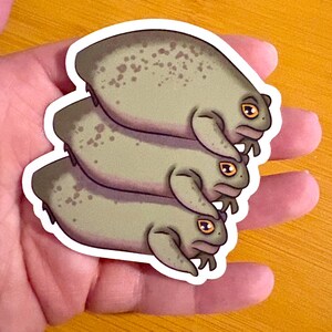 Puede incluir: Una pegatina con tres ranas de dibujos animados apiladas una encima de la otra. Las ranas son de color verde apagado con manchas verdes más oscuras y ojos amarillos. La pegatina tiene un borde blanco.