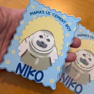 May include: Two blue and yellow stickers with a cartoon seal illustration. The stickers have the text "Mama's Lil' Cement Boy" and the name "Niko". The seal is white and gray with a smiling face.