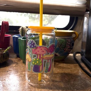 May include: A clear glass tumbler with a yellow lid and a matching yellow straw. The exterior features a colorful decal of various mushrooms in shades of purple, pink, green, and yellow, accented with small, raised, circular embellishments.