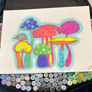 Peut inclure: Illustration colorée de plusieurs champignons stylisés dans les tons de rouge, rose, jaune, vert et violet, soulignés en noir. L'œuvre est sur papier blanc, avec un fond bleu clair. Une collection de marqueurs d'art est visible en bas.
