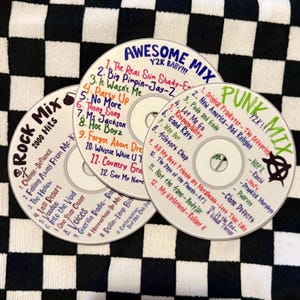 May include: Three silver compact discs with handwritten tracklists in colorful ink. The discs are labeled Awesome Mix Y2K Baby, Punk Mix Y2K, and Rock Mix 2000 Hits, featuring various song titles and band names written in red, blue, green, and black ink.