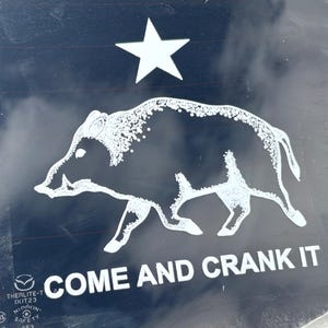 May include: White graphic of a boar and a star on a dark background. The text "COME AND CRANK IT" is below the boar. The image is on a car window.