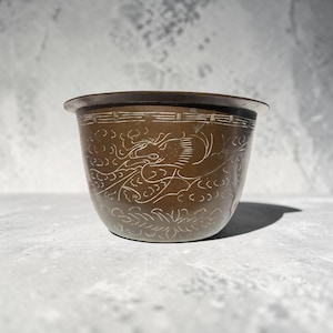 May include: A small, bronze-coloured planter with a detailed dragon design etched into the surface. The planter has a slightly flared rim and a decorative band near the top. The dragon design is visible on the front of the planter.