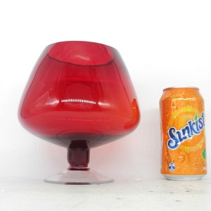 May include: A large, red glass bowl with a wide base and a rounded, flared top. The bowl is sitting on a white surface next to a can of Sunkist orange soda.