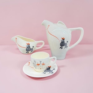 May include: A white ceramic tea set comprising a creamer, a teapot, and a teacup with a saucer. Each piece features a grey poodle illustration with an orange bow and a thin orange line. The teacup and saucer also display a house graphic.