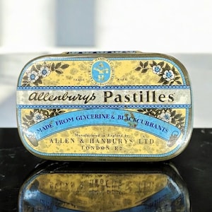 May include: A vintage Allenburys Pastilles tin, rectangular with rounded edges. The tin has a yellow background, blue and white floral details, and text reading "Allenburys Pastilles" and "Made from Glycerine & Blackcurrants". Made in London.