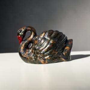 May include: A ceramic swan figurine featuring a glossy black finish with distressed copper-coloured accents throughout its body. The swan has a curved neck, detailed moulded wing feathers, and a bright red beak, presented in a side profile view.