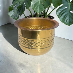 May include: A gold-coloured metal planter with a wide rim and a textured band of small, evenly spaced indentations. The planter is round and sits on a light-coloured surface, with a large green plant in the background.