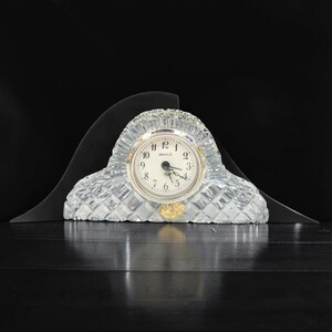May include: A clear, faceted glass mantel clock with a rounded arch shape. The clock face is white with black Arabic numerals and the text "INWALD" printed in the centre. A small gold-coloured sticker is attached to the lower front of the glass.