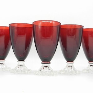 May include: Five ruby red wine glasses. Each glass has a conical bowl and sits atop a clear, textured base. The glasses are arranged in a row, showcasing their elegant design and rich colour. Perfect for a formal dinner setting.