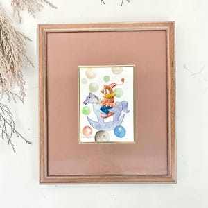 May include: A framed watercolour painting featuring a clown with a red hat and yellow collar riding a light blue rocking horse. The scene is decorated with colourful floating bubbles in shades of green, blue, and orange, all set within a wooden frame.