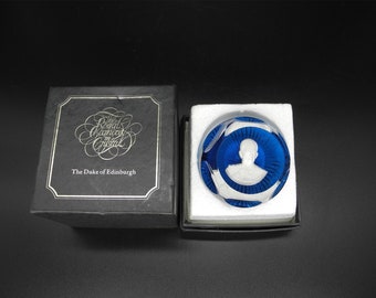 May include: A blue crystal paperweight with a white silhouette of a person inside. The paperweight is in a black box with the text "The Royal Family in Crystal" and "The Duke of Edinburgh".