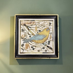 May include: A square ceramic tile framed in black with gold trim, featuring a detailed illustration of a yellow and blue bird perched on a branch. The text "GREENFINCH" is printed at the top, surrounded by black line drawings of berries and catkins.