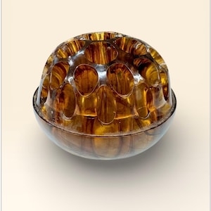 May include: An antique amber glass bowl with a distinctive pattern. The upper section has multiple oval apertures, and the base is a smooth, rounded shape. The glass has a glossy finish, reflecting light and creating a warm aesthetic. The bowl is likely from the mid-20th century.