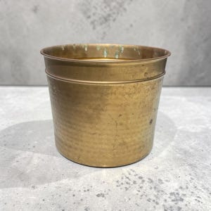 May include: A small, cylindrical brass planter with a hammered texture. The planter has a slightly flared rim and a tarnished, aged appearance, suggesting it is vintage or antique. Ideal for small plants or succulents.