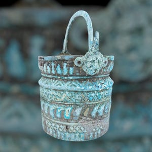 May include: A small, weathered, turquoise metal bucket with a curved handle. The bucket has intricate embossed patterns and a decorative element on the side. The design suggests an antique or vintage aesthetic, with a worn appearance.