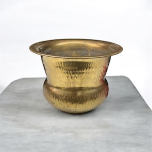 May include: A hammered brass bowl with a wide, rounded rim and a bulbous base. The bowl has a textured surface and a warm, golden colour. The bowl is sitting on a grey surface.