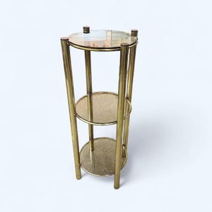 May include: A three-tiered plant stand with a gold metal frame. The stand features three circular shelves and a clear glass top. The stand is approximately 76 cm tall.