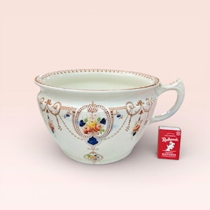 May include: A white ceramic bowl with a handle, decorated with floral and scroll patterns in blue, yellow, and orange. A small red matchbox is next to the bowl.
