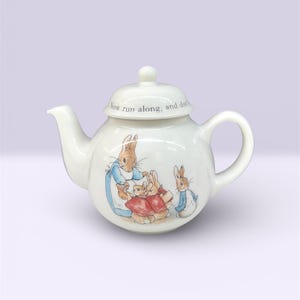 May include: White ceramic teapot with a lid, featuring a storybook illustration of rabbits. The teapot has a curved spout, a loop handle, and the text "Now run along, and don't" on the lid. A classic design for tea time.