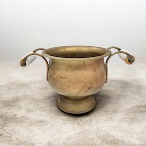 May include: A vintage brass bowl with two curved handles, each with a white ceramic grip. The bowl has a pedestal base and a slightly tarnished finish, indicating age and use. The handles are attached to the bowl with small metal brackets.