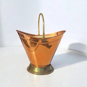 May include: A copper bucket with a brass handle and base. The bucket has a flared, trapezoidal shape with a decorative dotted line around the top. The handle is arched, and the base is a contrasting brass colour.