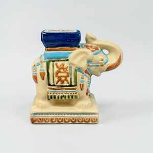 May include: A ceramic elephant figurine with a blue seat and decorative accents in turquoise, brown, and green. The elephant has a cream-coloured body and is standing on a rectangular base.