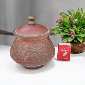 May include: A maroon-coloured, lidded jar with embossed floral patterns. The jar has a brass-coloured knob on the lid. A small box of Redheads matches sits next to the jar, and a small potted plant with pink flowers is in the background.