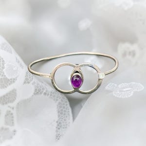 May include: A silver-toned metal bangle bracelet featuring a central design of two interlocking circular loops. A single, smooth, oval-shaped purple gemstone is set vertically within the centre of the overlapping circles of the metal band.