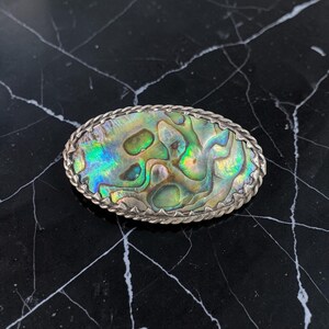 May include: An oval abalone shell brooch with a silver-tone rope border. The iridescent shell features green, blue, and gold colours. The brooch is set against a black marble surface.