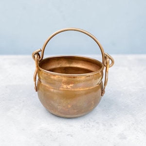 May include: A small, antique-style copper cauldron with a rounded body and a curved handle. The cauldron has a tarnished, aged appearance, suggesting it is vintage. The handle is attached to the sides with decorative loops.