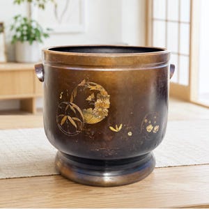 May include: A cylindrical bronze planter with a dark, weathered patina and a wide, flared base. The exterior features gold-toned inlaid designs of bamboo stalks, floral arrangements, and small circular motifs, complemented by two small side handles.