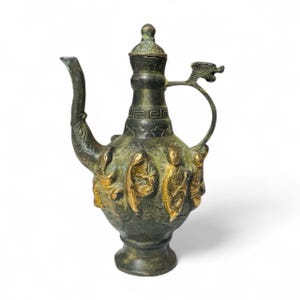 May include: An antique bronze-coloured pitcher with a dragon-shaped handle and lid. The body of the pitcher features gold-coloured relief figures. The spout is curved, and the pitcher sits on a small base. The design suggests an Asian influence.