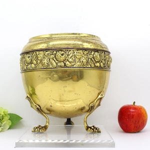 May include: A gold-toned brass planter with a floral design and three legs. The planter is on a clear acrylic base.