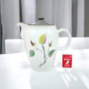 May include: White ceramic teapot with a grey lid and handle, decorated with a floral design in green, yellow, and red. A small red matchbox with white text is next to the teapot. The teapot has a spout and a curved handle.