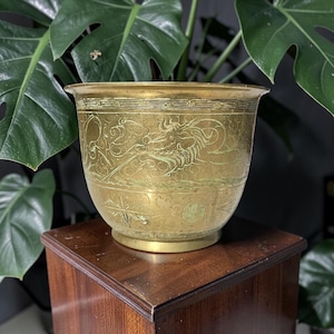 May include: A brass planter featuring an engraved dragon motif encircling the exterior. The vessel has a wide, flared rim and a circular base, with intricate linear patterns and geometric symbols etched into the metallic surface of the pot.
