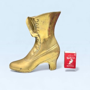May include: A gold-coloured brass boot ornament with a high heel and button detailing. The boot is upright. A small box of Redheads safety matches is next to the boot.