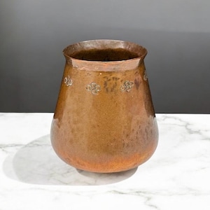 May include: A copper-coloured vase with a wide base and a narrow opening. The vase has a textured surface and is adorned with small, embossed floral designs. The vase is likely used for decorative purposes.