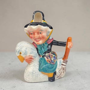 May include: A ceramic creamer shaped like a figure wearing a black hat and teal dress, riding a white goose with a yellow beak. The figure holds a brown broom handle, and the piece features a glossy finish with hand-painted details in various colours.