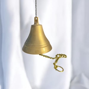 May include: A brass bell with a gold chain and a small ring. The bell is suspended from a black chain. The bell has a classic shape and is made of shiny, polished brass. The background is a white fabric.