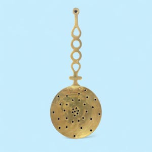 May include: A vintage brass tea infuser. It has a circular, perforated bowl and a decorative handle. The handle has teardrop-shaped cutouts and a loop at the top. The bowl has a central pattern of small holes. A tea accessory.